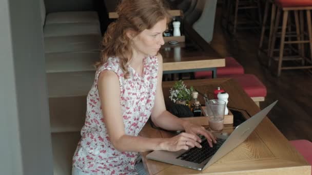 Une jeune femme d'affaires est assise à une table dans un restaurant. Elle portait un ordinateur portable avec accès à Internet, il vérifie le courrier et boire du café. Elle a l'air très heureuse — Video