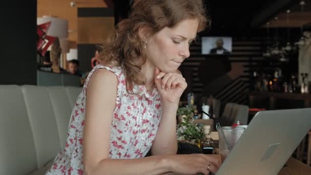 En ung forretningskvinne sitter ved et bord på en restaurant. Hun hadde en bærbar pc med tilgang til Internett, den sjekker posten og drikker kaffe. Hun ser veldig lykkelig ut. – stockvideo