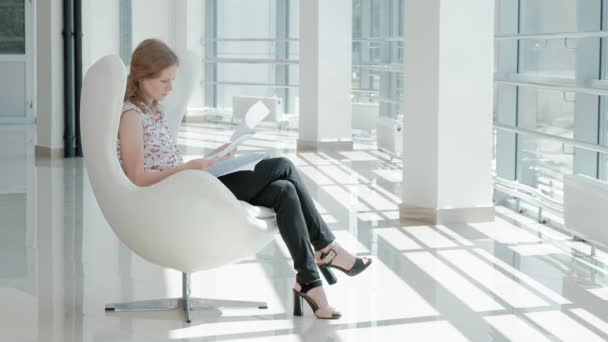 ガラスのオフィスとチェックのドキュメントの白い椅子に座って魅力的な実業家 — ストック動画