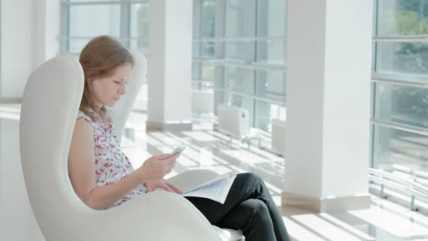Atractiva mujer de negocios sentada en una silla blanca en una oficina de vidrio y comprueba documentos — Vídeos de Stock