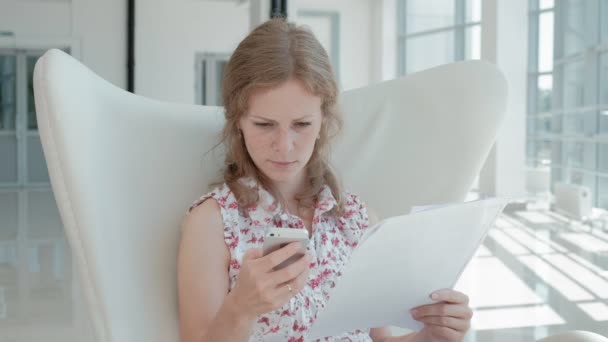 Attraente donna d'affari seduta su una sedia bianca in un ufficio di vetro e controlla i documenti — Video Stock
