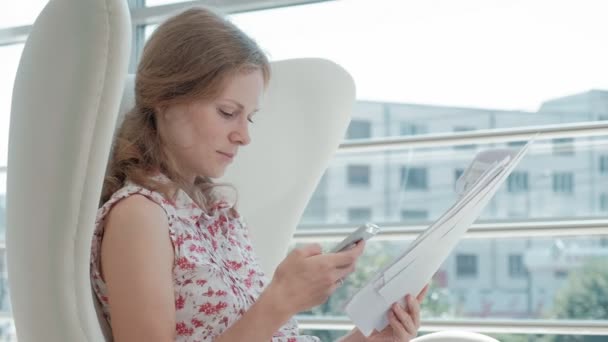 Attractive businesswoman sitting on a white chair in a glass office and checks documents — Stock Video