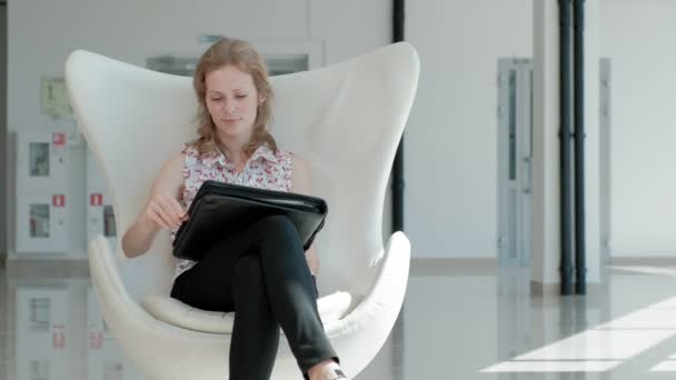 Attraktive Geschäftsfrau sitzt auf einem weißen Stuhl in einem gläsernen Büro und prüft Dokumente — Stockvideo