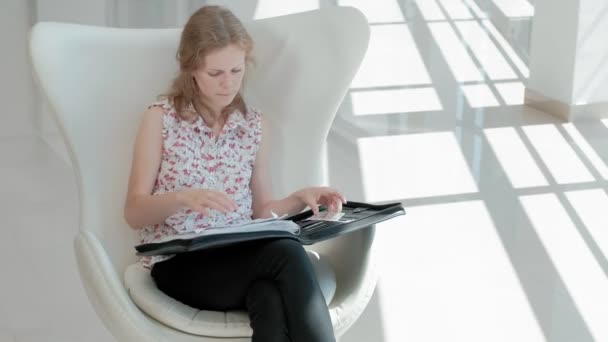 Attraktive Geschäftsfrau sitzt auf einem weißen Stuhl in einem gläsernen Büro und prüft Dokumente — Stockvideo