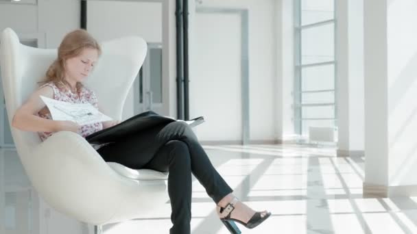 Ελκυστικός επιχειρηματίας που κάθεται σε μια λευκή καρέκλα σε ένα γυάλινο γραφείο και ελέγχους έγγραφα — Αρχείο Βίντεο
