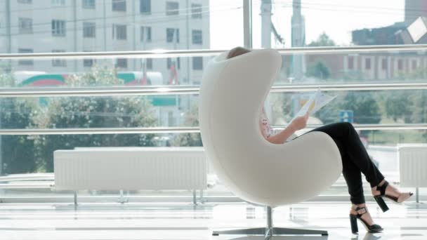 Привлекательная деловая женщина сидит на белом стуле в стеклянном кабинете и проверяет документы — стоковое видео