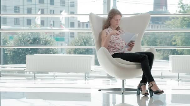 Atractiva mujer de negocios sentada en una silla blanca en una oficina de vidrio y comprueba documentos — Vídeo de stock