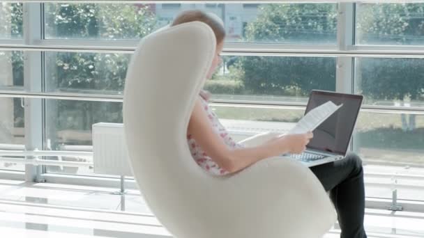 Jolie femme d'affaires assise sur une chaise blanche dans un bureau en verre et travaillant derrière un ordinateur portable — Video