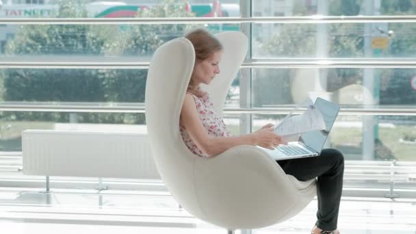 Atractiva mujer de negocios sentada en silla blanca en la oficina de cristal y trabajando detrás de la computadora portátil — Vídeos de Stock