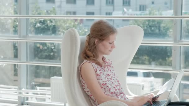 Attractive businesswoman sitting on white chair in glass office and working behind laptop — Stock Video