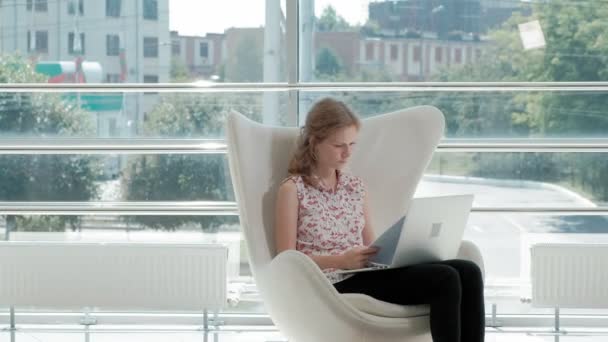 Приваблива бізнес-леді сидить на білому стільці в скляному офісі і працює позаду ноутбука — стокове відео