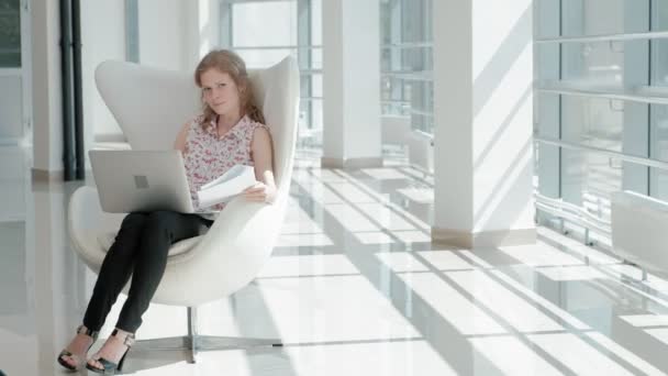 Aantrekkelijke zakenvrouw zittend op een witte stoel in glas kantoor en werken achter een laptop — Stockvideo