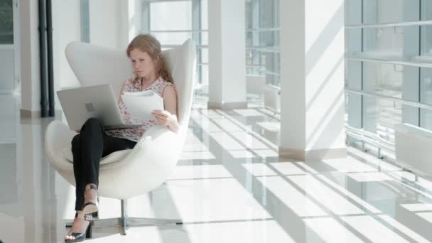 Aantrekkelijke zakenvrouw zittend op een witte stoel in glas kantoor en werken achter een laptop — Stockvideo