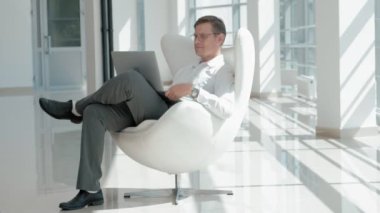 Bir cam ofis beyaz bir sandalyede oturan ve bir dizüstü bilgisayar üzerinde çalışan olgun işadamı