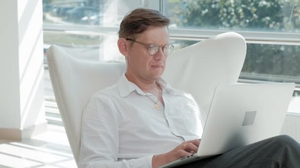 Rijpe zakenman zittend op een witte stoel in een glas kantoor en werken op een laptop — Stockvideo