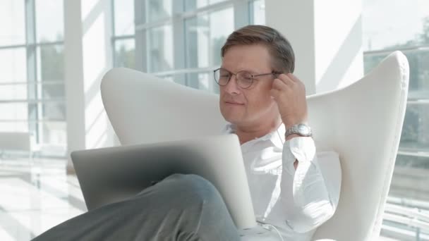Hombre de negocios maduro sentado en una silla blanca en una oficina de vidrio y trabajando en un ordenador portátil — Vídeo de stock