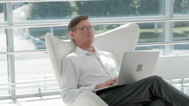 Mature businessman sitting on a white chair in a glass office and working on a laptop — Stock Video