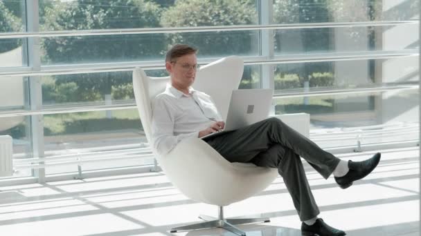 Homem de negócios maduro sentado em uma cadeira branca em um escritório de vidro e trabalhando em um laptop — Vídeo de Stock