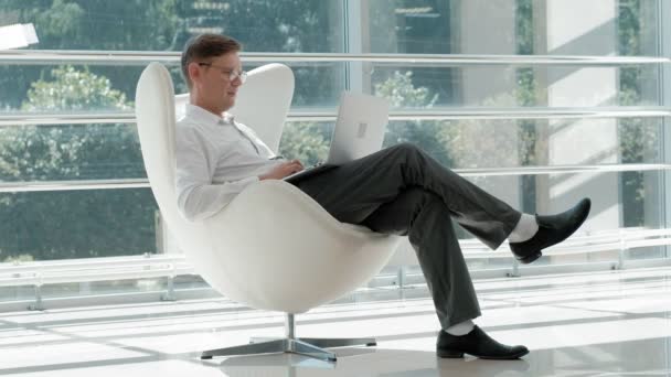 Kypsä liikemies istuu valkoisella tuolilla lasitoimistossa ja työskentelee kannettavan tietokoneen — kuvapankkivideo