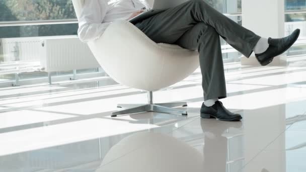 Mature businessman sitting on a white chair in a glass office and working on a laptop — Stock Video