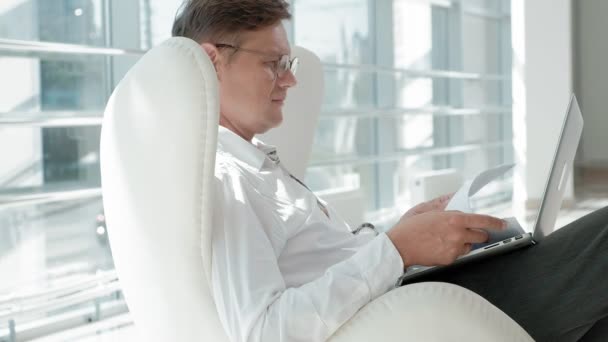 成熟したビジネスマン ガラス オフィスの白い椅子に座って、ノート パソコンに取り組んで — ストック動画