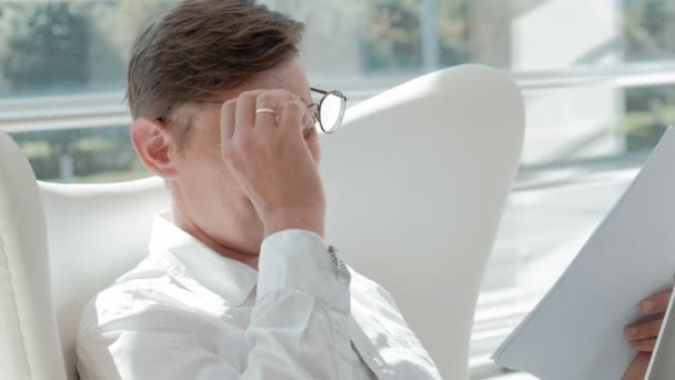 Vonzó üzletember ül egy fehér széket, egy üveg-office és ellenőrzések dokumentumokban — Stock videók