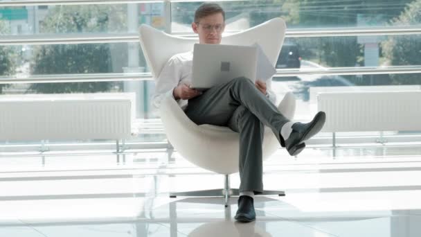 Зрелый бизнесмен, сидящий на белом стуле в стеклянном офисе и работающий над ноутбуком — стоковое видео