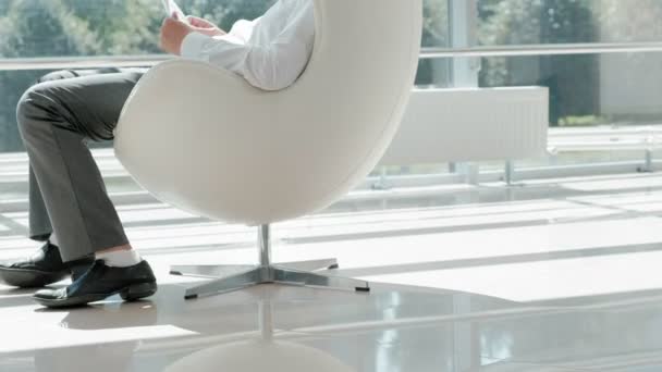 Hombre de negocios maduro sentado en una silla blanca en una oficina de vidrio y trabajando en un ordenador portátil — Vídeos de Stock