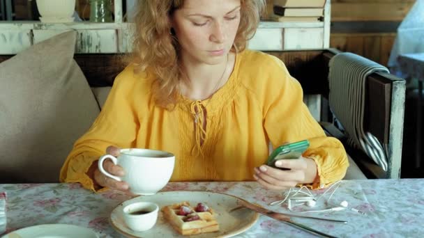 Hermosa mujer está comiendo gofres en la cafetería utiliza un teléfono inteligente para tomar una foto — Vídeo de stock