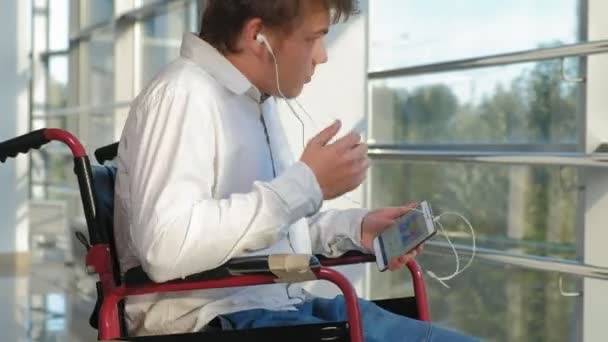 Man op een rolstoel op een venster luisteren naar muziek op hoofdtelefoons van een smartphone uitgeschakeld — Stockvideo