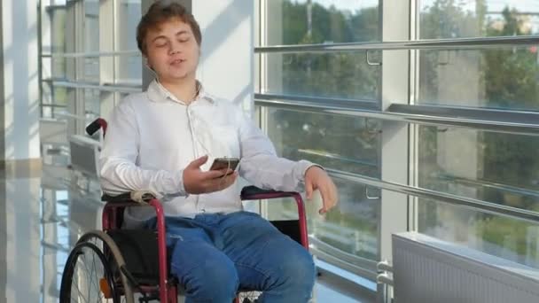 Hombre discapacitado en una silla de ruedas en una ventana escuchando música en auriculares desde un teléfono inteligente — Vídeos de Stock