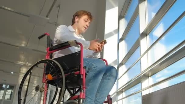 Инвалид на инвалидной коляске у окна слушает музыку на наушниках со смартфона — стоковое видео