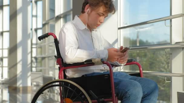 Uomo disabile su una sedia a rotelle ad una finestra che ascolta musica sulle cuffie da uno smartphone — Video Stock