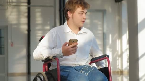 Инвалид на инвалидной коляске у окна слушает музыку на наушниках со смартфона — стоковое видео
