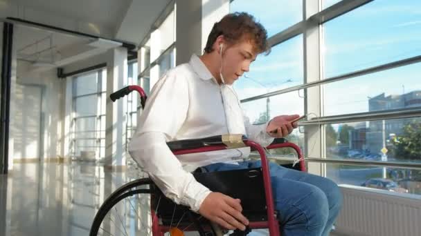 Hombre discapacitado en una silla de ruedas en una ventana escuchando música en auriculares desde un teléfono inteligente — Vídeo de stock