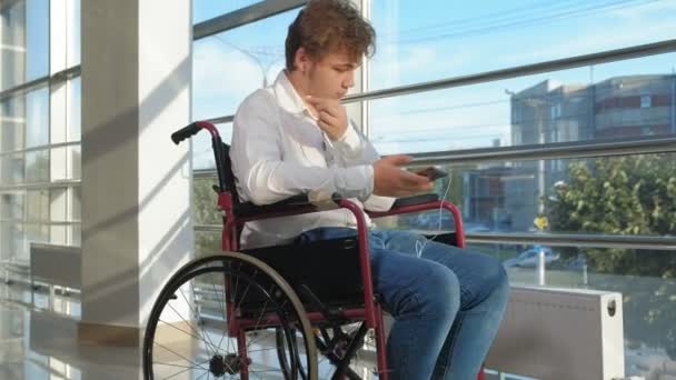 Hombre discapacitado en una silla de ruedas en una ventana escuchando música en auriculares desde un teléfono inteligente — Vídeo de stock