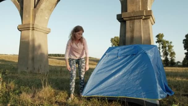 Giovane donna raccoglie una tenda turistica vicino al vecchio ponte — Video Stock