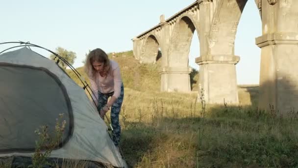 Молодая женщина собирает туристическую палатку возле старого моста — стоковое видео