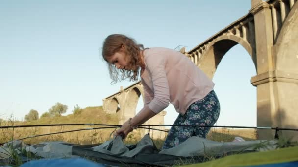 Jonge vrouw verzamelt een toeristische tent in de buurt van de oude brug — Stockvideo