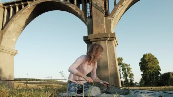 Giovane donna raccoglie una tenda turistica vicino al vecchio ponte — Video Stock