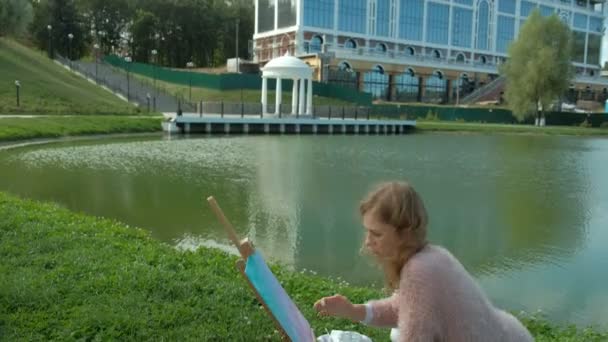 Een mooie vrouw met rood haar, schetst een beeld op doek, dat op de ezel staat. De dame is in de open lucht in de buurt van het meer van de rivier, ze tekent uit het leven — Stockvideo