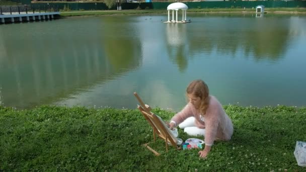 Красивая женщина с рыжими волосами, рисует картину на холсте, который стоит на мольберте. The lady is in the open air near the lake of the river, she draw from life — стоковое видео