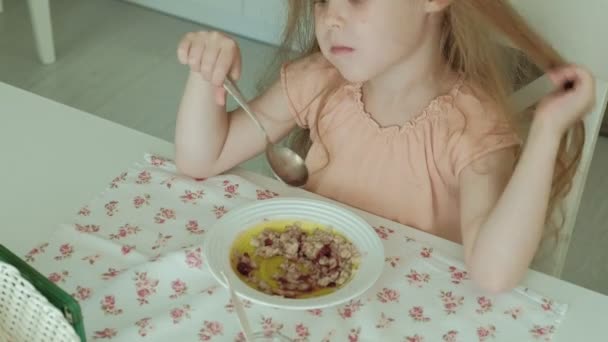 Happy beautiful girl eating porridge for breakfast and having fun in white kitchen at home — Stock Video