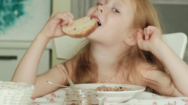 Heureuse belle fille manger du porridge pour le petit déjeuner et s'amuser dans la cuisine blanche à la maison — Video