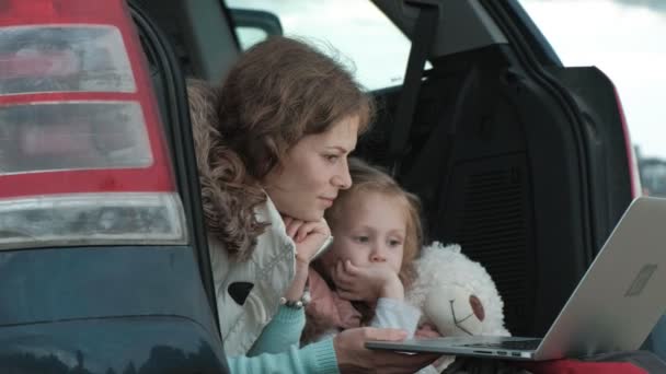 Vacker ung kvinna och hennes lilla dotter sitter i öppna bagageluckan på en bil på flodstranden av havet njuter en laptop — Stockvideo