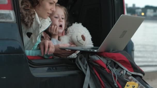 Mooie jonge vrouw en haar dochtertje zitten in de open kofferbak van een auto op de oever van de zee genieten van een laptop — Stockvideo