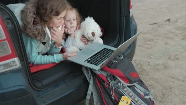 Hermosa joven y su hija pequeña están sentados en el maletero abierto de un coche en la orilla del río del mar disfrutando de un ordenador portátil — Vídeos de Stock