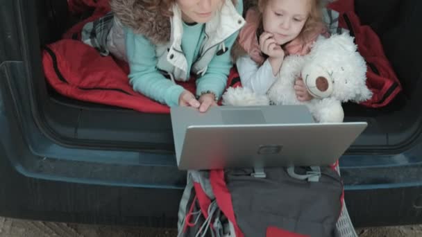 Красивая молодая женщина и ее маленькая дочь сидят в открытом багажнике автомобиля на берегу моря наслаждаясь ноутбуком — стоковое видео