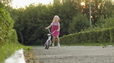 Uzun sarı saç bir pembe etek ve jumper mutlu, güzel, küçük bir kızla yolda o gülümsüyor bir çocuk bisiklet sürmek. Çok yavaş hareket