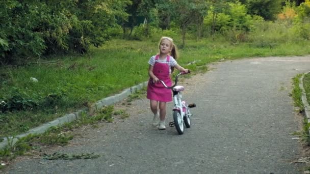 Uma menina feliz, bonita, com longos cabelos loiros em uma saia rosa e jumper monta uma bicicleta infantil na estrada, ela sorri. Movimento super lento — Vídeo de Stock